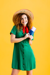 happy woman in straw hat holding passport with oar ticket isolated on yellow