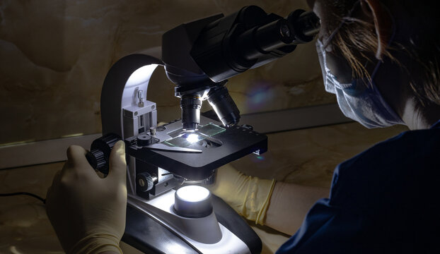 Photo of a laboratory assistant sitting in a dark room and working with a light microscope. The lab assistant is wearing a medical mask and silicone gloves.