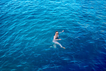 Cute caucasian adult girl in sunglasses swims in blue water - top view. Young woman holds on to the turquoise-blue surface of the sea or pool on a summer sunny day