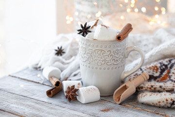 Obraz na płótnie Canvas Winter hot drink. Cozy home composition with white mug with chocolate and marshmallow, cinnamon. Knitted mittens, christmas lights, wooden background. Festive holiday atmosphere, family spirit. Close
