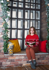 A blonde girl in a red sweater sits on the windowsill.