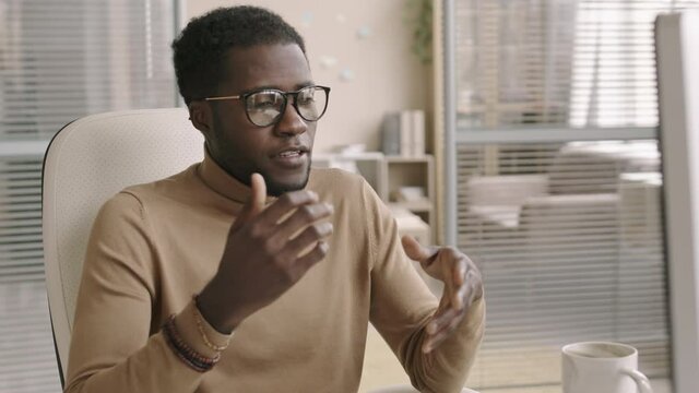 Medium shot of young African businessman wearing casual clothes and eyeglasses sitting at workplace in office and talking to investor distantly using computer