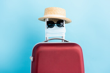 sunglasses, medical mask and straw hat on red luggage on blue