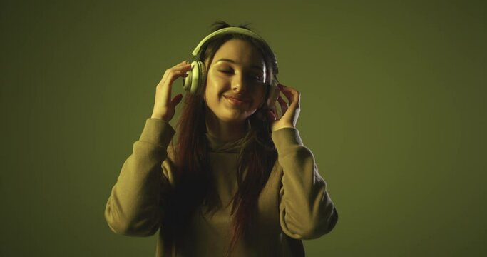 Carefree energetic young woman with ponytail, wearing hoodie, listening music in headphones, dancing actively, smiling. Colourful flashing background.
