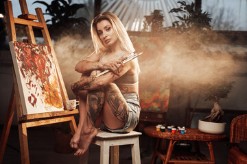 Obraz na płótnie Canvas Cuddling legs seductive woman with blond hairs poses on chairs holding paintbrushes in dark atmospheric room with smoke.