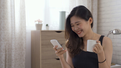 portrait of pretty asian japanese lady having morning coffee and watching funny videos on phone online while relaxing in bed.