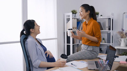 cheerful asian expectant woman laughing and chatting with her business partner while having hot coffee in paper cup in modern office.