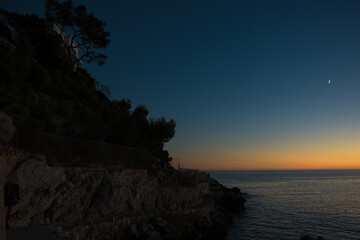 crépuscule sur la mer avec un dégradé de couleurs du bleu foncé à l'orange dans le ciel, un croissant de lune et le littoral dans la pénombre à gauche de l'image 