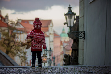 Beautiful toddler child with lantern and teddy bear, casually dressed, looking at night view of Prague city