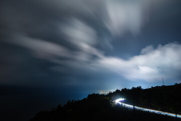 Night landscape. Curved road with traces of headlights on moving clouds background