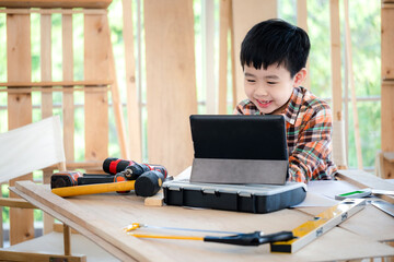 Young Asia boy wearing a shirt and sitting at the table at home and see the tablet. Many equipment tools (hammer, screwdriver, saw) on the table. Carpenter and education concept. Smile and happy