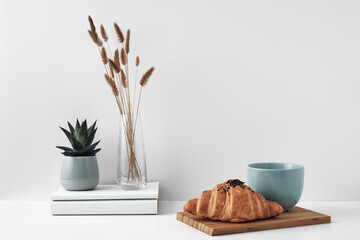 Croissant and a mug on a wooden stand, a vase and a pot with a plant on the books, white background. Eco-friendly materials in interior decor, minimalism. Copy space, mock up.