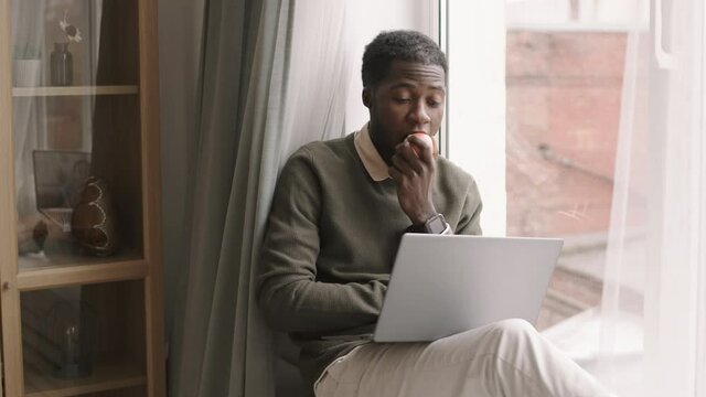Lockdown of young African man wearing casual clothes sitting on windowsill, watching video using laptop while eating apple