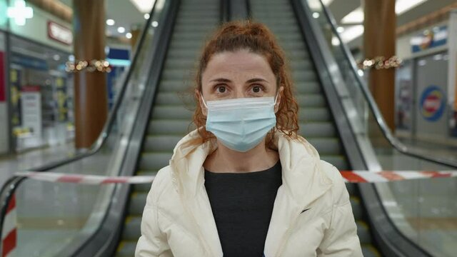 Young woman wearing covid-19 mask stands in front an escalator with barrier tape in huge mall look in camera, locked in lockdown. Shopping during coronavirus in mall. Pandemic, virus, danger, epidemic