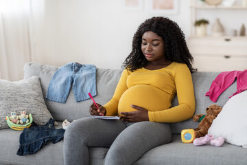 Concentrated pregnant woman sitting on sofa with notebook and pen