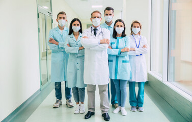 Serious confident doctors team in safety medical masks with crossed arms are looking on the camera on hospital background