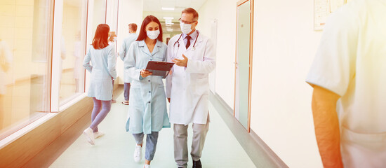 Two confident doctors in safety medical masks have a discussion about some treatment methods during walking the hospital corridor with other colleagues around them