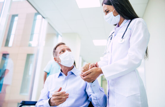 Ill Senior Man With Protective Safety Mask On Face In A Wheelchair And A Confident Doctor In The Medical Mask While Transporting On The Hospital.