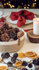 Obraz na płótnie Canvas Festive holiday table vintage style with pine cones and dried fruit as decoration, a red napkin and a white table cloth in front of a decorated Christmas tree with lights