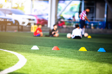 Obraz premium Selective focus to cone marker is soccer training equipment on green artificial turf with blurry kid players training background. Material for training class of football academy.