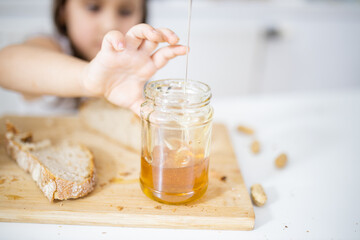 Little girl picking honey from a jar next to a slice of bread