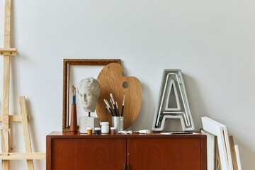 Stylish composition of artist workspace room with design retro teak commode, poster frames, easel, book, decoration and painting accessories. Template.
