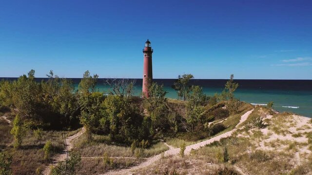 Beautiful aerial view panning in towards the Little Sable Lighthouse along the shoreline of Lake Michigan sitting amongst vegetation and tree covered sand dunes with a clear blue sky above.