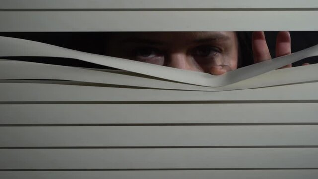 Close up front portrait from indoors of brunette woman pushing blinds at window with hands. Looking fearfully outside