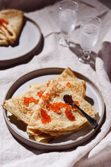 Traditional Russian crepes or pancakes with red caviar on gray plates and glasses of ice vodka over greige linen tablecloth. Pancake week or Shrovetide