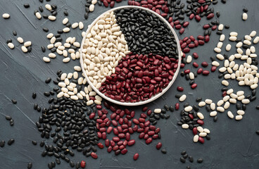 beans in white ceramic plate. bowl of beans. black bean, red kidney bean and white bean. navy bean, cannellini bean, white kidney bean on dark gray background and scattered various beans