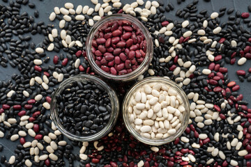 glass jar with black bean, red kidney bean and white bean. navy bean, cannelini bean, white kidney bean on  dark gray background and scattered various beans. triangle shape made from beans jar
