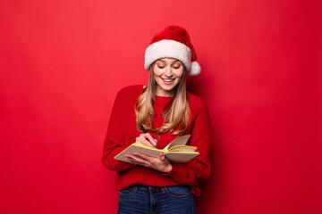 Young woman with Santa hat holds a notepad in her hands and bites a pen checking the list of gifts or children with good behavior isolated on a red background.,