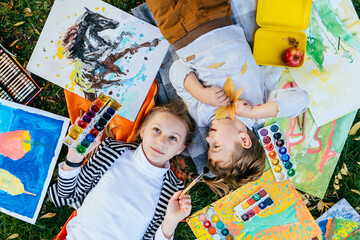 Outdoor portrait of pretty girl with her smaller brother lying on fresh green grass with paints,...