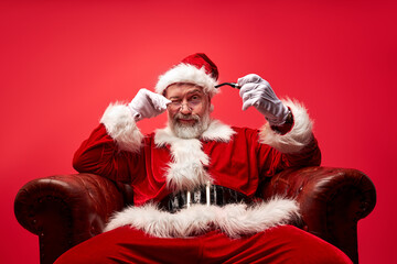 arrogant santa sitting on chair isolated on red background, caucasian senior elderly male in santa costume has big abdomen and bad character