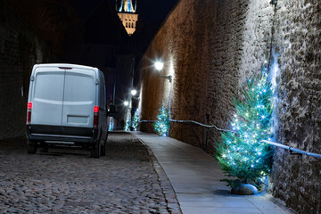 A white cargo van delivering presents in a narrow medieval cobblestone city street with illuminated...