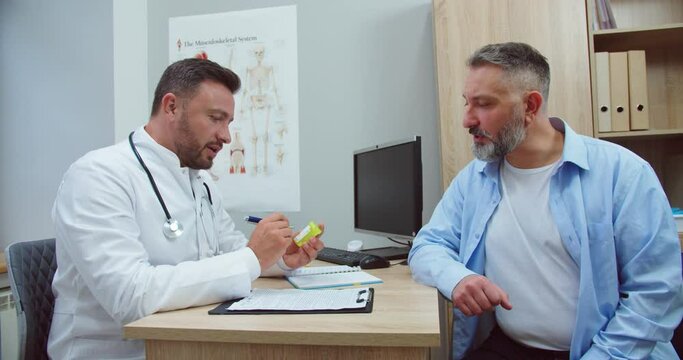 Side view. Portrait of handsome Caucasian focused male doctor speaking and consulting middle-aged man patient prescribing medicine while sitting in hospital cabinet. Treatment concept