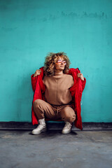 Young fashionable woman with curly hair, in red jacket crouching in front of the wall and looking away.