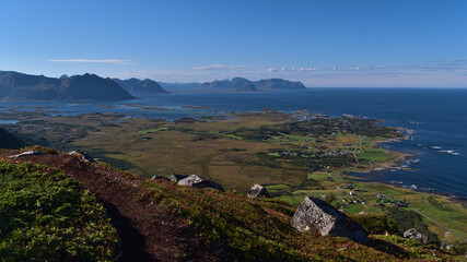 Beautiful panoramic view over northern coast of Lofoten islands, Norway with village Straumnes, blue Norwegian Sea, rugged mountains and hiking path through meadows in foreground on sunny summer day.