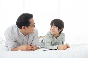 Father and Asian boy writing on book,lying on bed at home, Dad and son spending quality time together.