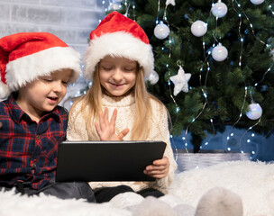 Fototapeta na wymiar children in santaclaus hats sit near a Christmas tree on a white rug with a tablet in their hands, video call with relatives on the eve of New Year's celebrations