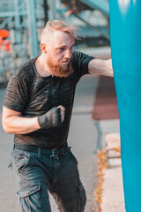 Fototapeta na wymiar Street Fighter in Black Clothes and Bandages on the Wrist Boxing in Punching Bag Outdoors. Young Man Doing Box Training and Practicing His Punches at the Outside Gym