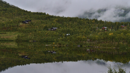 Traditional wooden houses surrounded by green forest on the shore of Lapphaugvatnet lake in northern Norway reflected in the calm water on cloudy day in late summer.