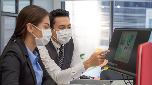 Two analysts with face masks discussing financial data on computer.