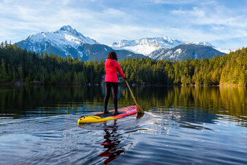 Adventurous Girl Paddle Boarding on Levette Lake with famous Tantalus Mountain Range in the background. Taken in Squamish, North of Vancouver, British Columbia, Canada.