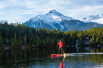 Fototapeta na wymiar Adventurous Girl Paddle Boarding on Levette Lake with famous Tantalus Mountain Range in the background. Taken in Squamish, North of Vancouver, British Columbia, Canada.