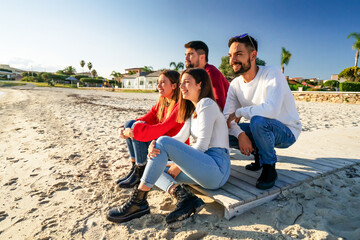 Fototapeta premium A group of happy young friends traveling in nature bent over and sitting on a wooden boardwalk at the beach smiling while watching the sunset or sunrise on their winter beach vacation as a couple