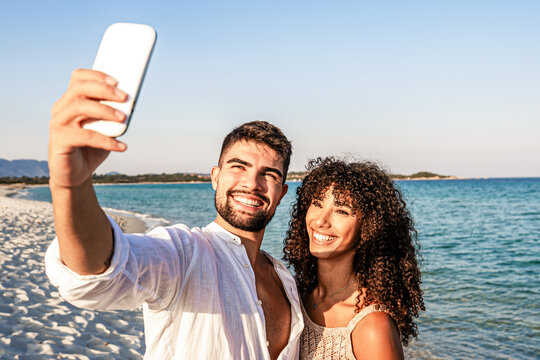 Beautiful young mixed-race couple in love smiling looking to the phone for a self portrait at sunset or dawn on beach with sea water in background - Handsome guy makes a toothy selfie with girlfriend