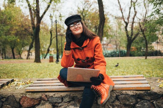 Young smiling woman in a coat and hat with a laptop sitting on a bench