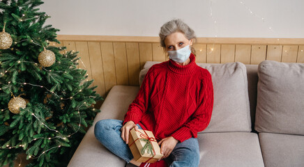 Sad senior woman wearing protective mask sitting on couch alone with a gift in Christmas decorated room . Celebrating Christmas home during the coronavirus pandemic concept.
