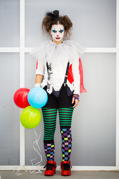 A standing woman dressed up as a terrorific and devilish clown in many colors and a colorful makeup. The woman has three balloons in her hands. Perfect for Halloween or a scary themed party.